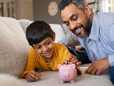 Happy indian son saving money in piggy bank with father. Lovely ethnic father teaching to little boy importance of saving money for future. Smiling middle eastern kid adding coin in piggybank while lying on couch with dad at home.