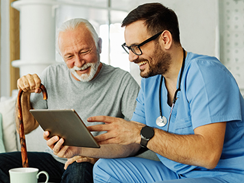 Doctor or nurse caregiver showing a tablet screen to  senior man and laughing at home or nursing home