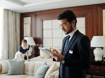 Hotel manager inside a hotel room on his iPad while a worker fixes the bed