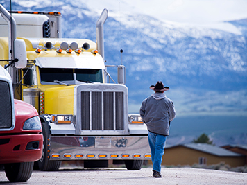 The truck driver goes to his bright yellow attractive impressive customized big rig semi truck parked in the parking lot on a picturesque backdrop of snow-capped mountain ranges.